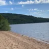 Praia tranquila com areia dourada e vegetação ao fundo, destacando a beleza natural da paisagem à beira do lago sob um céu azul.