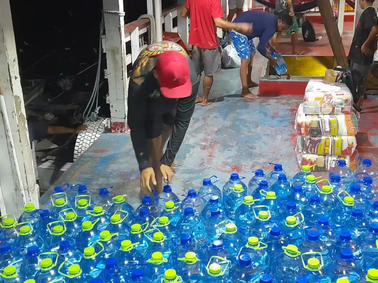 Homens descarregando garrafas de água em um barco, organizando a carga durante a noite. Um ambiente movimentado com diversos tripulantes.