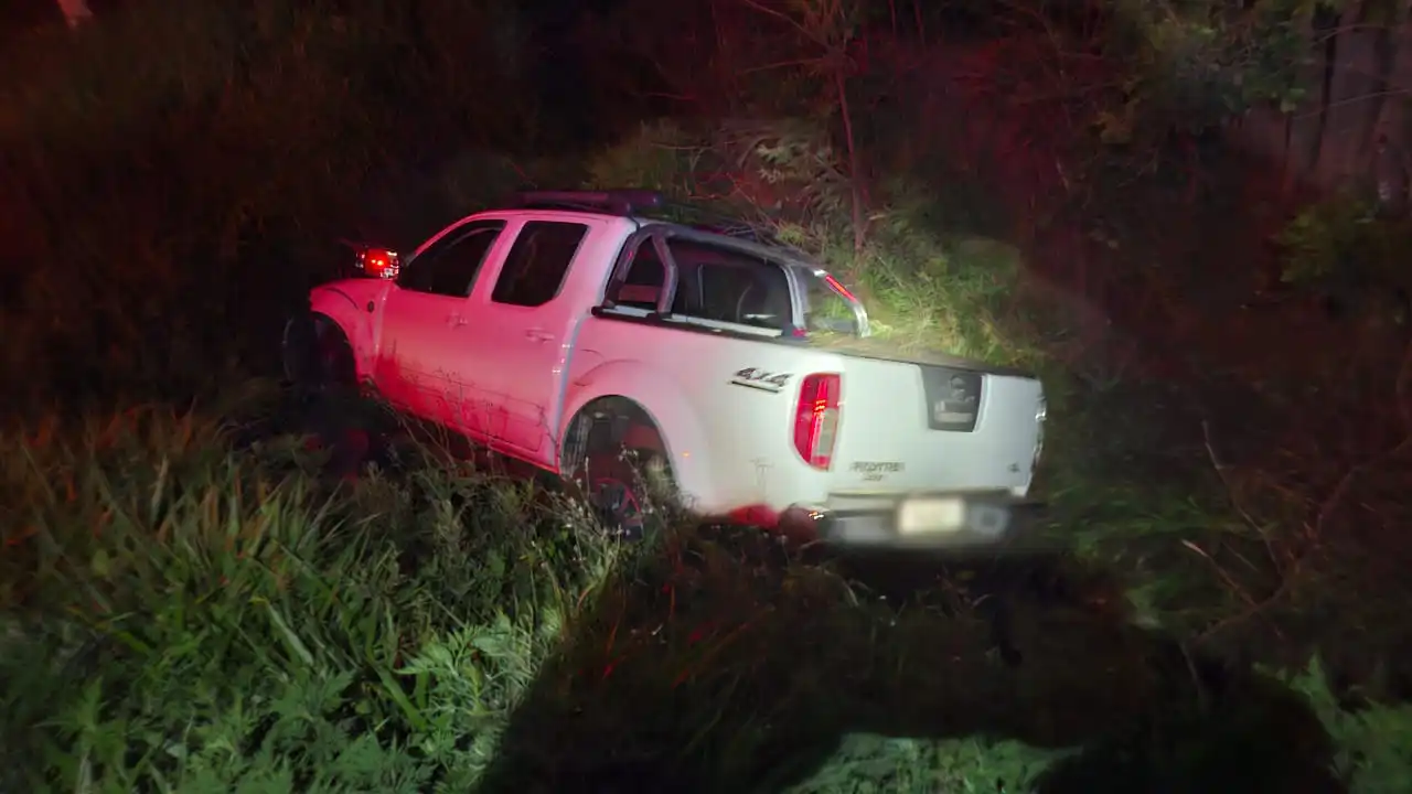 Imagem do acidente na ERS-324, em Passo Fundo, onde uma caminhonete branca está fora da pista, cercada por vegetação, com luzes de emergência.