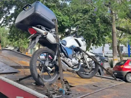 Imagem de uma motocicleta sendo rebocada por um guincho, destacando sua cor branca e azul, e o cenário urbano ao fundo.