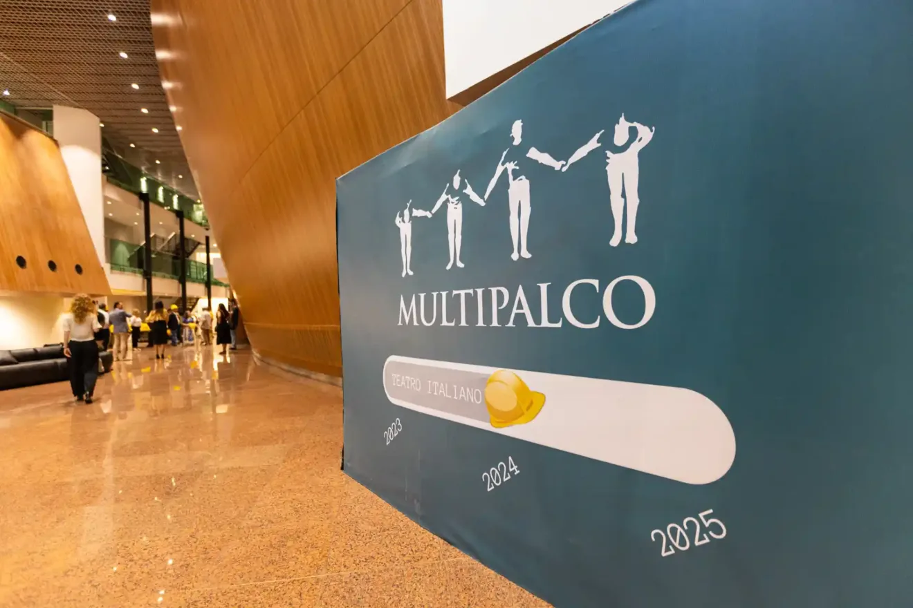 Imagem da inscrição "Multipalco Teatro Italiano" em um ambiente de evento. O banner destaca as datas 2023, 2024 e 2025, simbolizando a continuidade do projeto.