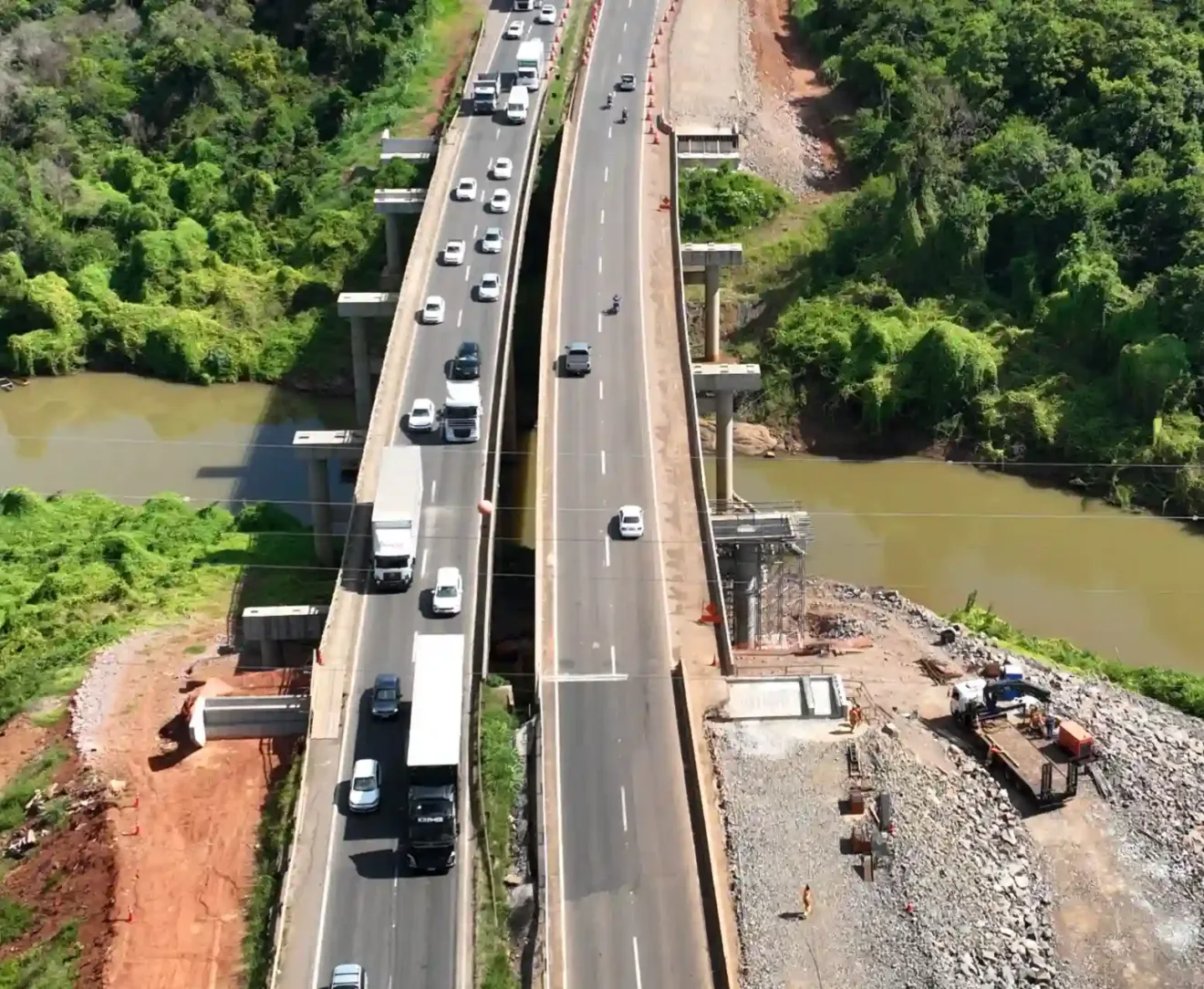 Vista aérea das obras na ponte do Arroio Boa Vista, com tráfego intenso de veículos, cercada por vegetação densa e um rio ao lado.