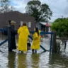 Imagem mostra pessoas em área alagada, usando capas de chuva amarelas, discutindo sobre a situação de inundação, com casas ao fundo.