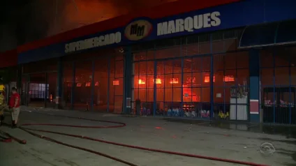 Incêndio no Supermercado Marques em Guaíba, com chamas visíveis pela fachada e equipe de bombeiros atuando na contenção do fogo.