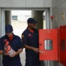 Dois bombeiros em atividade, um segurando um extintor e o outro abrindo um armário de emergência.