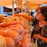 Mulher escolhendo batatas em feira livre, cercada por diversas variedades de batatas frescas em um mercado ao ar livre, com barraca colorida ao fundo.