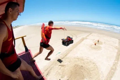 Salvamento aquático na praia, com dois salva-vidas em ação. Um salva-vidas salta da torre enquanto um quadriciclo está na areia.