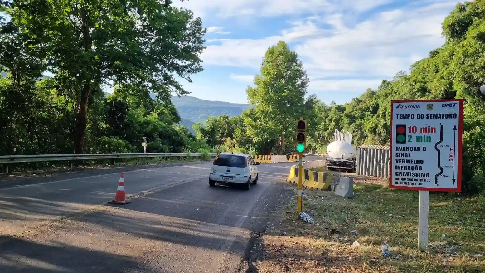 A imagem mostra um sinal de trânsito com um semáforo verde, indicando 10 minutos para avançar. O local é cercado por vegetação e uma estrada em condições variáveis, com cones de sinalização visíveis.