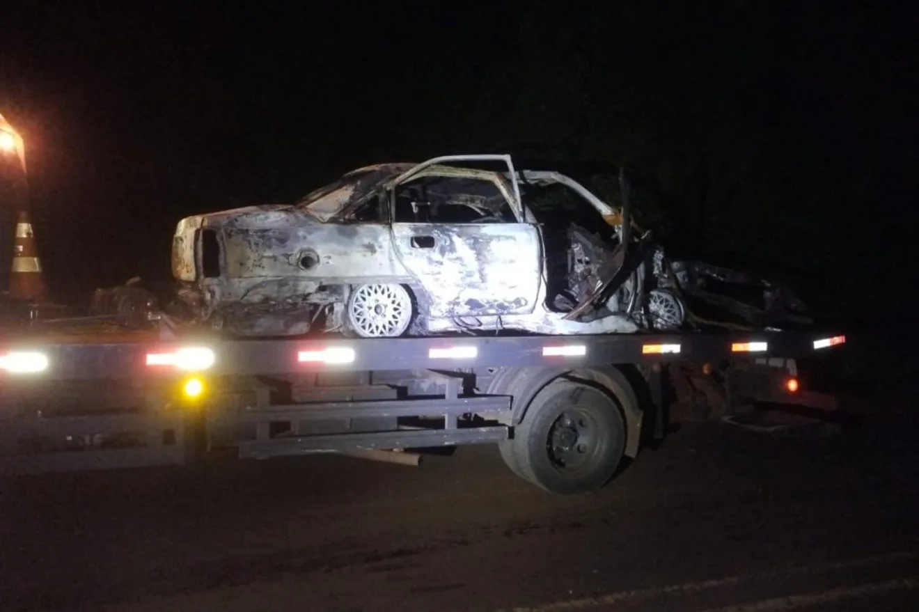 Imagem de um carro queimado sendo transportado em um caminhão durante a noite.