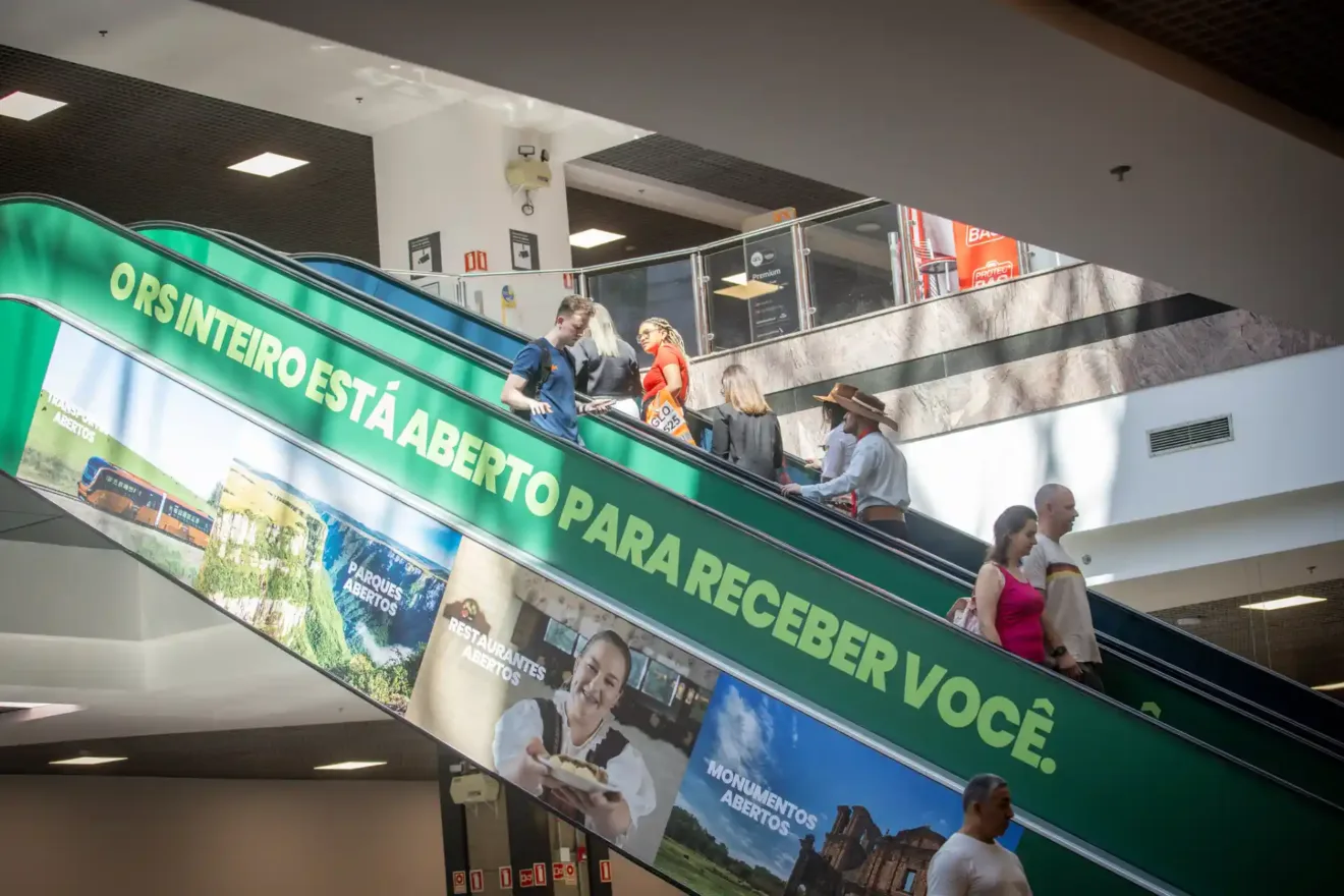 A imagem mostra pessoas descendo uma escada rolante no Aeroporto Salgado Filho, com uma faixa verde destacando que o RS inteiro está aberto para receber turistas.