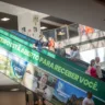 A imagem mostra pessoas descendo uma escada rolante no Aeroporto Salgado Filho, com uma faixa verde destacando que o RS inteiro está aberto para receber turistas.