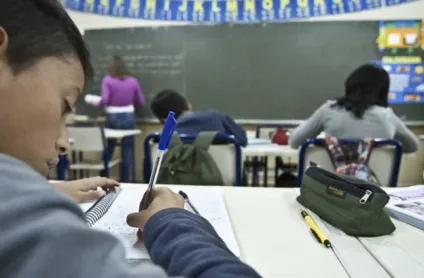 Crianças estudando em sala de aula, com foco em um aluno escrevendo em um caderno, demonstrando o ambiente de aprendizado.