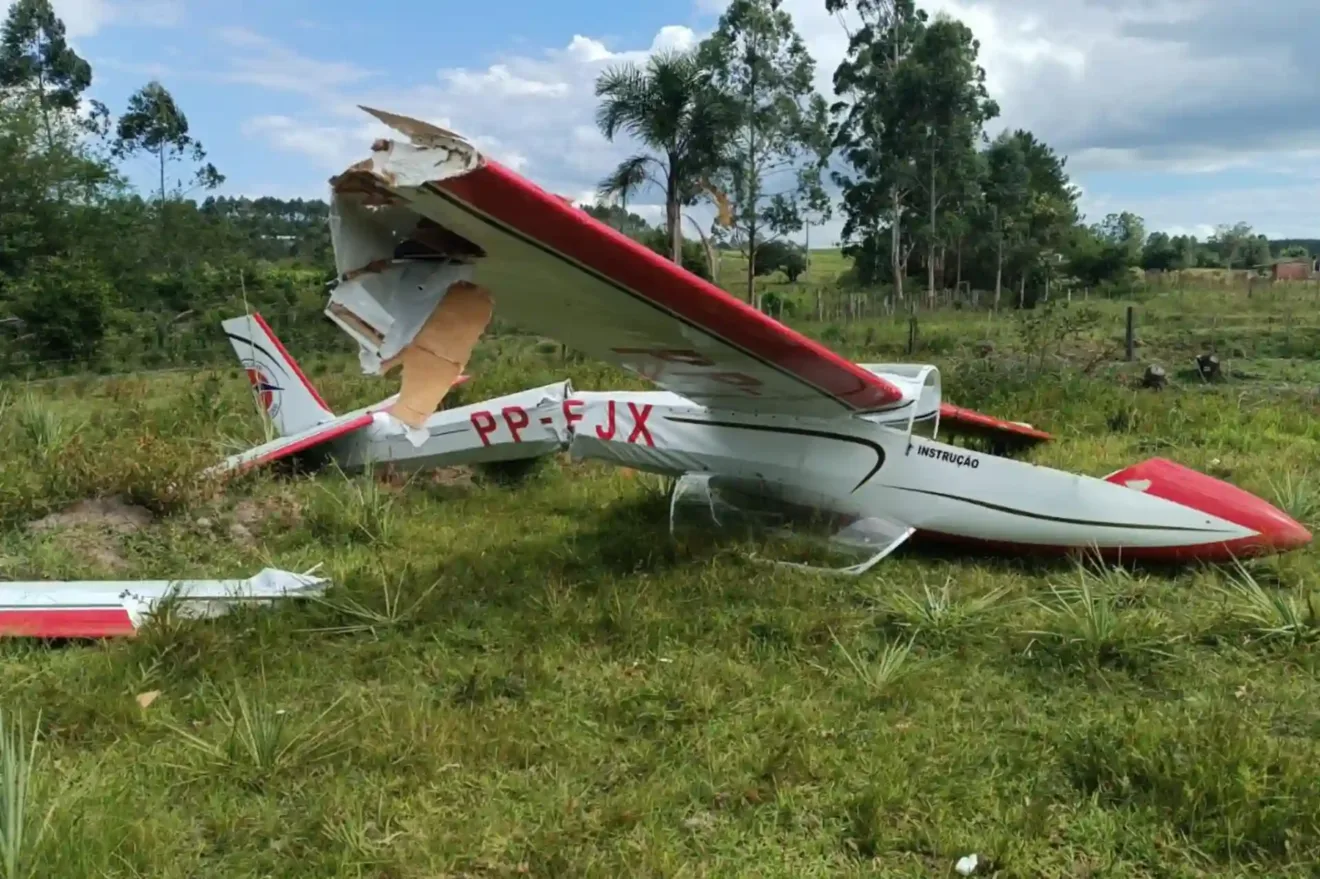 Imagem de um avião acidentado em uma área verde, com a fuselagem danificada e as asas quebradas. O cenário apresenta vegetação ao fundo, após um acidente aéreo em Montenegro.