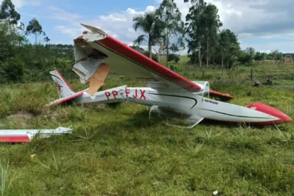 Imagem de um avião acidentado em uma área verde, com a fuselagem danificada e as asas quebradas. O cenário apresenta vegetação ao fundo, após um acidente aéreo em Montenegro.