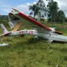 Imagem de um avião acidentado em uma área verde, com a fuselagem danificada e as asas quebradas. O cenário apresenta vegetação ao fundo, após um acidente aéreo em Montenegro.