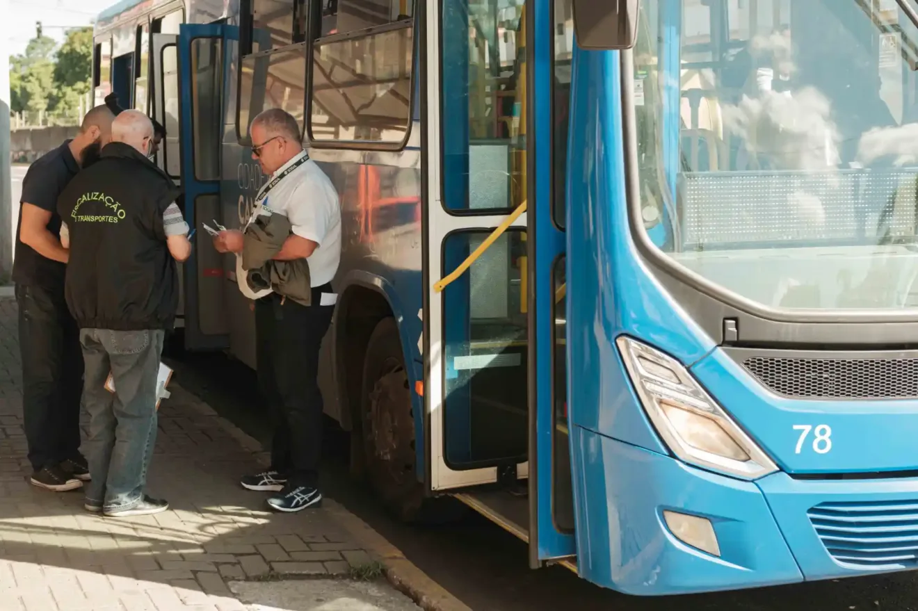 Três homens observam um ônibus azul parado, um deles verifica documentos enquanto os outros observam. O ônibus é parte do transporte público de Canoas, na região metropolitana.