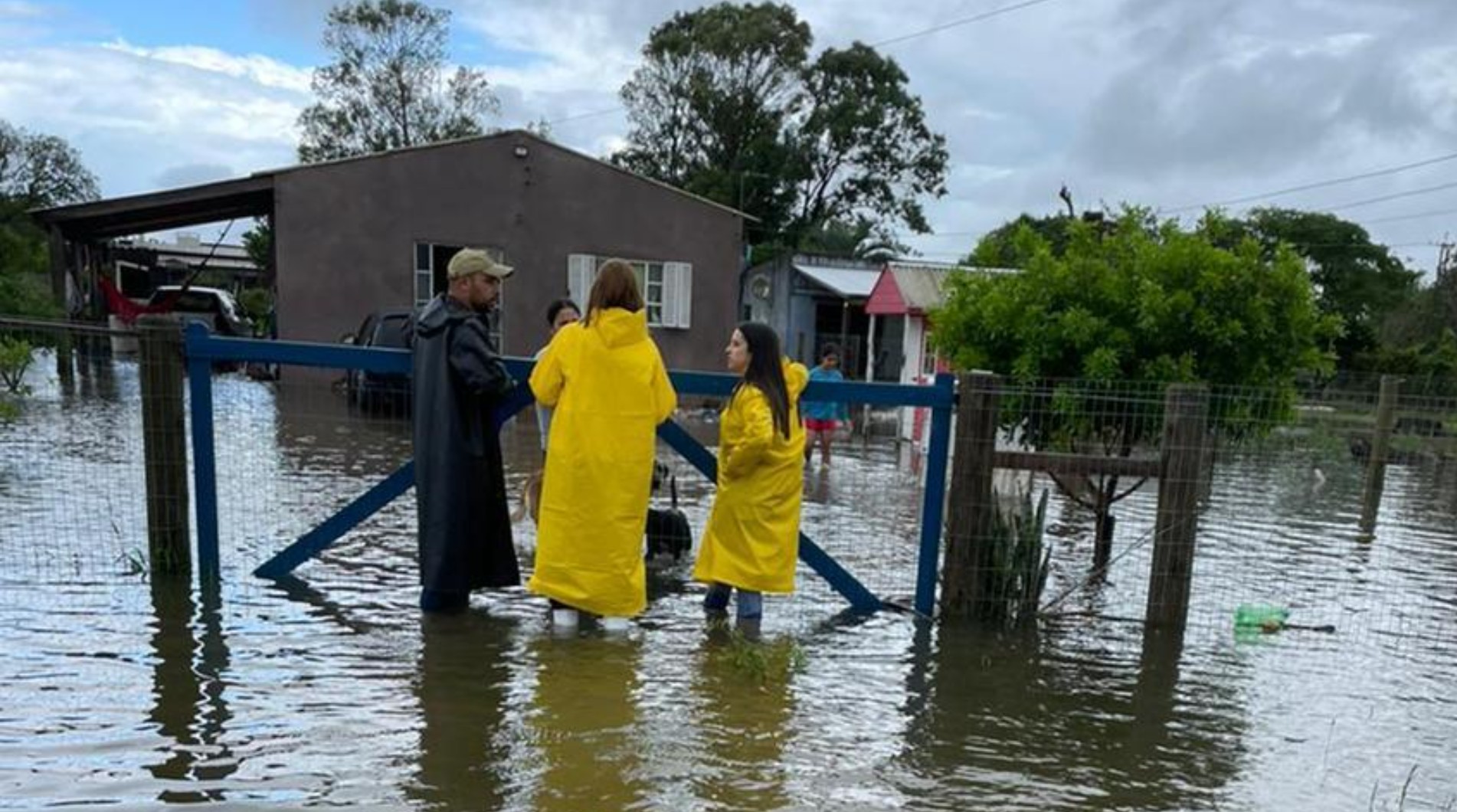 Imagem mostra pessoas em área alagada, usando capas de chuva amarelas, discutindo sobre a situação de inundação, com casas ao fundo.