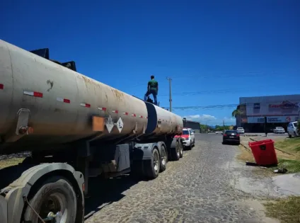 Um homem em cima de um caminhão-tanque em uma rua ensolarada. O caminhão possui tanque grande e é cercado por veículos.