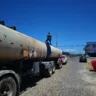 Um homem em cima de um caminhão-tanque em uma rua ensolarada. O caminhão possui tanque grande e é cercado por veículos.