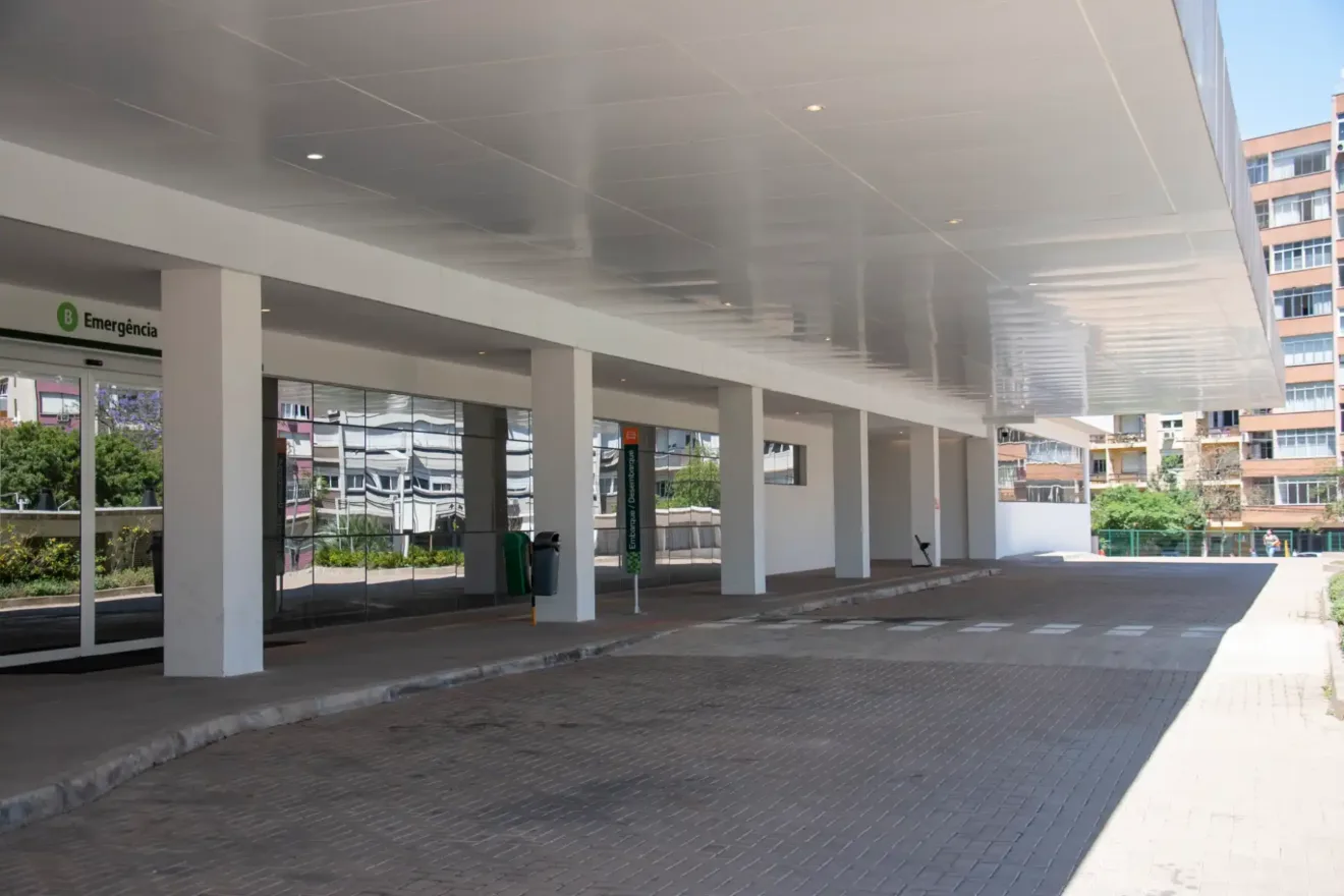 Foto da entrada da Emergência do Bloco B do Hospital de Clínicas de Porto Alegre, mostrando a área coberta e espaço livre. A estrutura é moderna e bem iluminada, rodeada por vegetação.