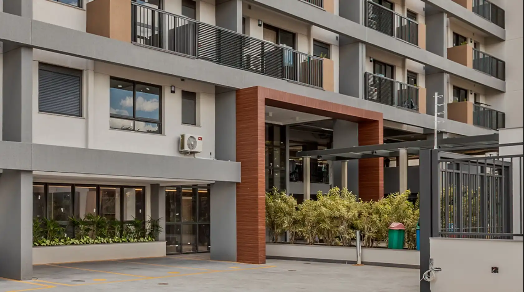 Imagem de fachada de um moderno edifício residencial com jardins e áreas externas, destacando detalhes arquitetônicos e a entrada principal.