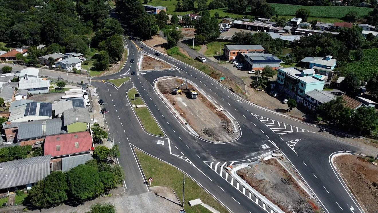 Vista aérea de uma rotatória em construção, rodeada por casas e áreas verdes, mostrando o fluxo de tráfego na cidade.