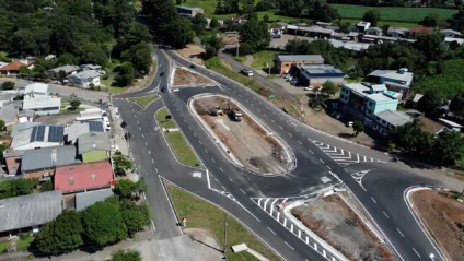 Vista aérea de uma rotatória em construção, rodeada por casas e áreas verdes, mostrando o fluxo de tráfego na cidade.