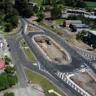 Vista aérea de uma rotatória em construção, rodeada por casas e áreas verdes, mostrando o fluxo de tráfego na cidade.