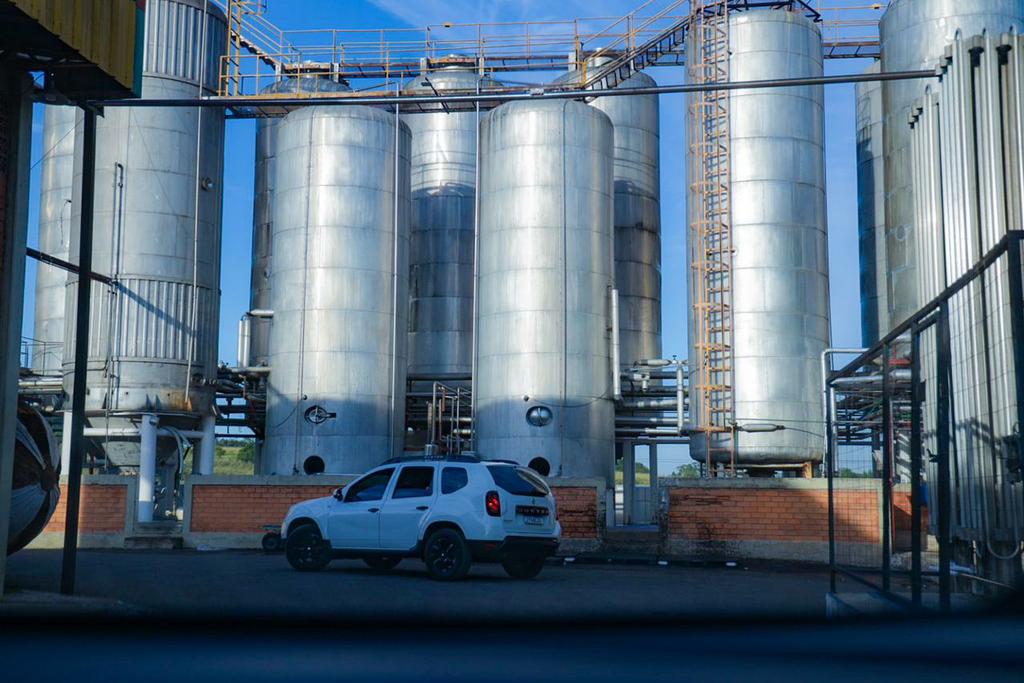 Vista de uma fábrica com silos metálicos e um carro branco estacionado em primeiro plano.