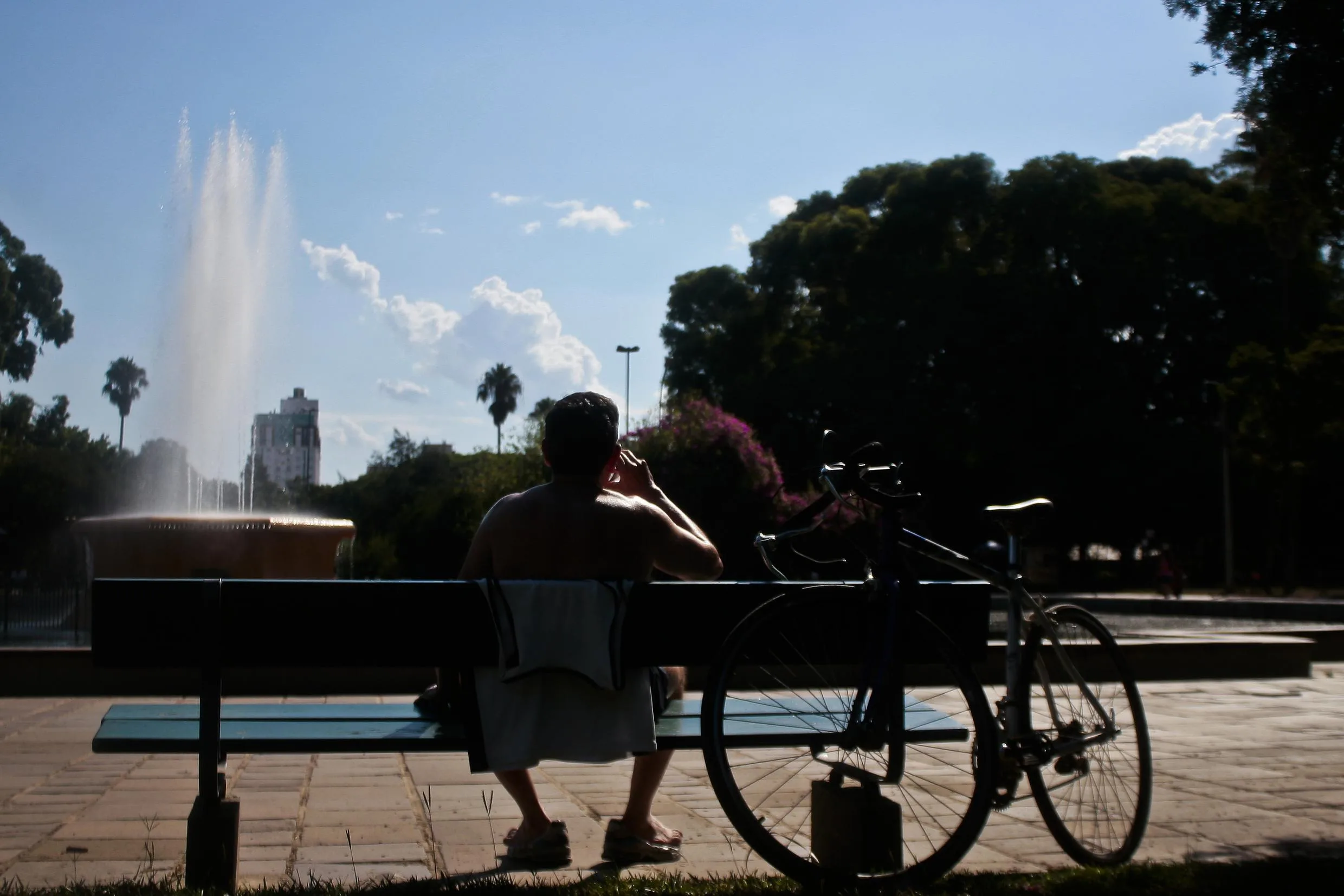 Pessoa relaxando em um banco de parque, com uma fonte ao fundo e uma bicicleta ao lado, aproveitando um dia ensolarado em Porto Alegre