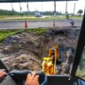 Imagem mostrando a operação de escavação com maquinário pesado, focando na profundidade da vala e no motorista na cabine do equipamento.