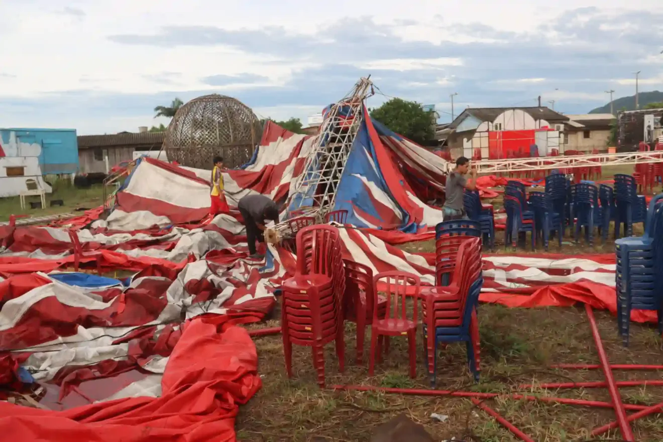 Imagem de um circo após uma tempestade, mostrando tendas rasgadas e cadeiras desordenadas. 