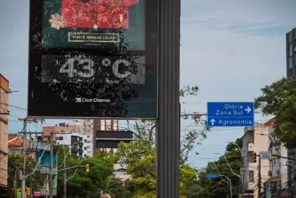 Imagem de um termômetro de rua mostrando a temperatura de 43°C e uma placa de sinalização indicando direções em uma cidade em dia ensolarado.