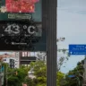 Imagem de um termômetro de rua mostrando a temperatura de 43°C e uma placa de sinalização indicando direções em uma cidade em dia ensolarado.