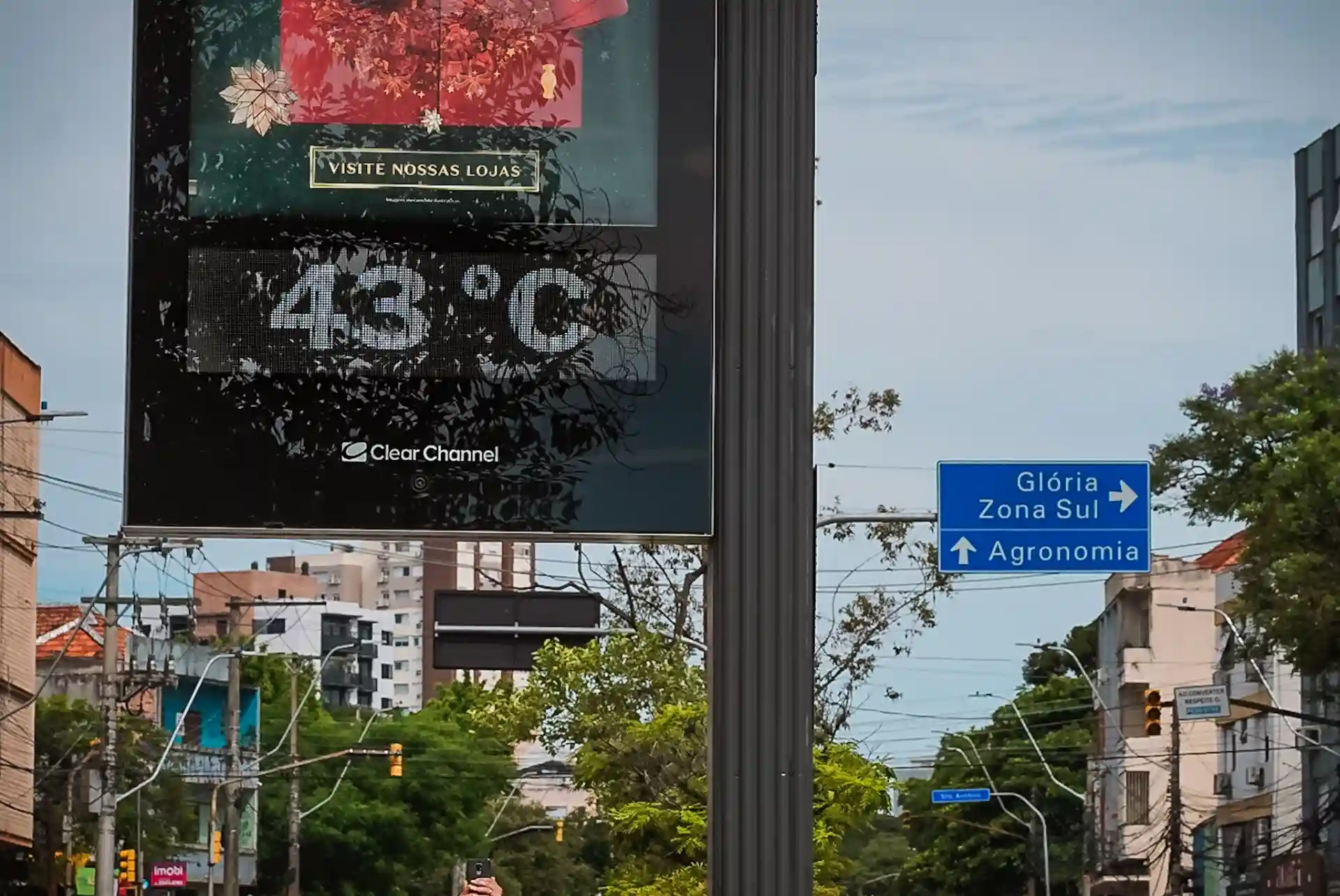 Imagem de um termômetro de rua mostrando a temperatura de 43°C e uma placa de sinalização indicando direções em uma cidade em dia ensolarado.