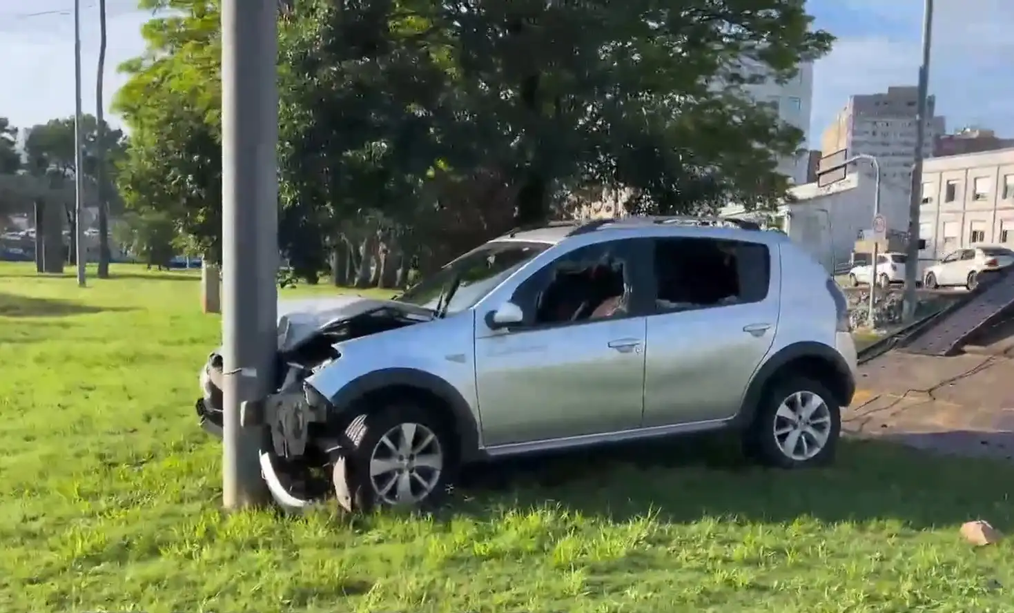 Imagem de um carro prata colidido com um poste em um campo verde. O carro apresenta danos significativos e janelas quebradas.