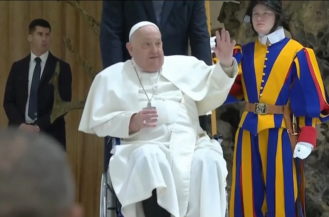 Papa Francisco acenando enquanto está em uma cadeira de rodas, ao lado de um guarda suíço em uniforme colorido, representando a liderança da Igreja Católica.