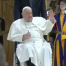 Papa Francisco acenando enquanto está em uma cadeira de rodas, ao lado de um guarda suíço em uniforme colorido, representando a liderança da Igreja Católica.