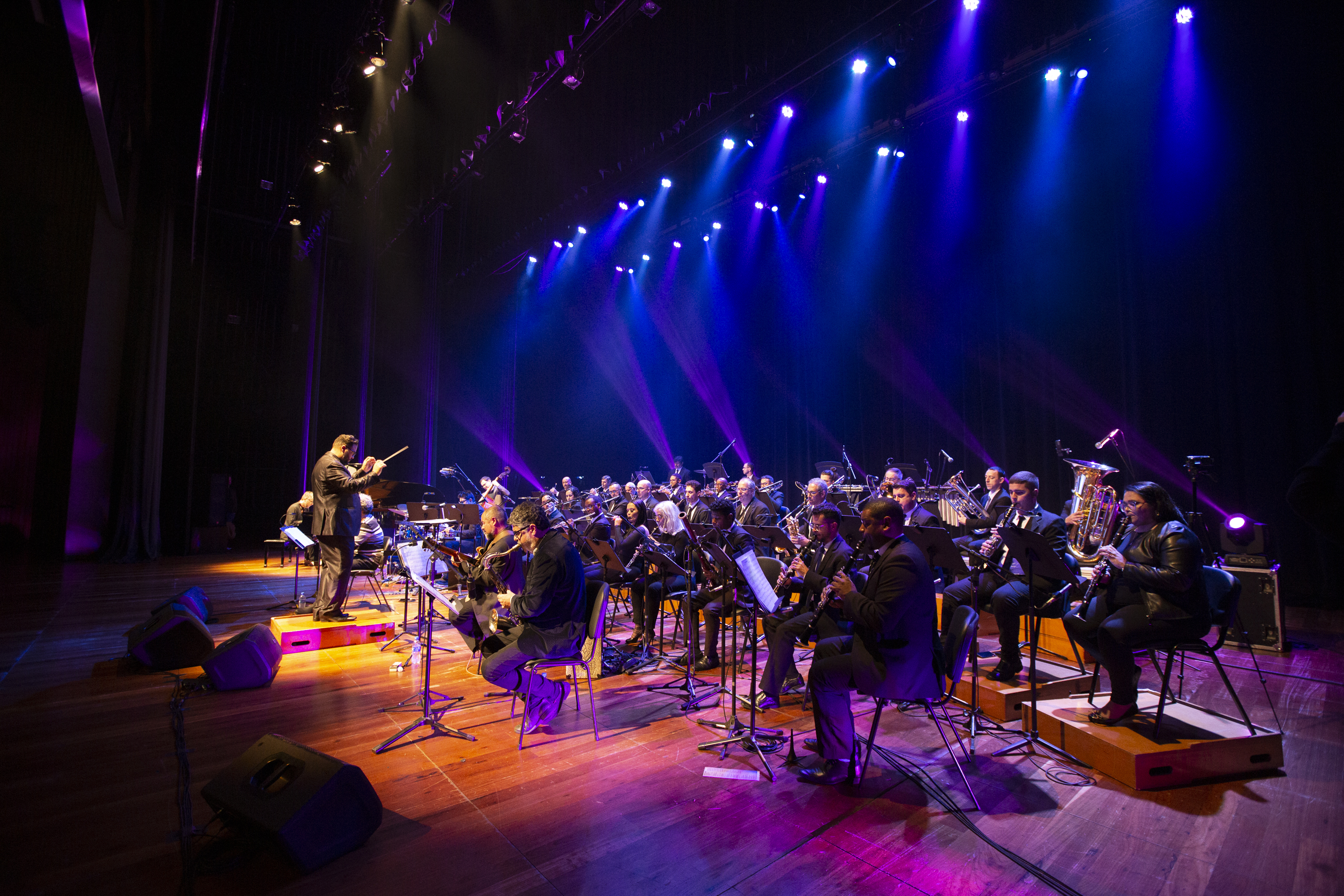 Imagem da Orquestra de Sopros de Novo Hamburgo em uma apresentação ao vivo, com músicos em um palco iluminado por luzes coloridas.