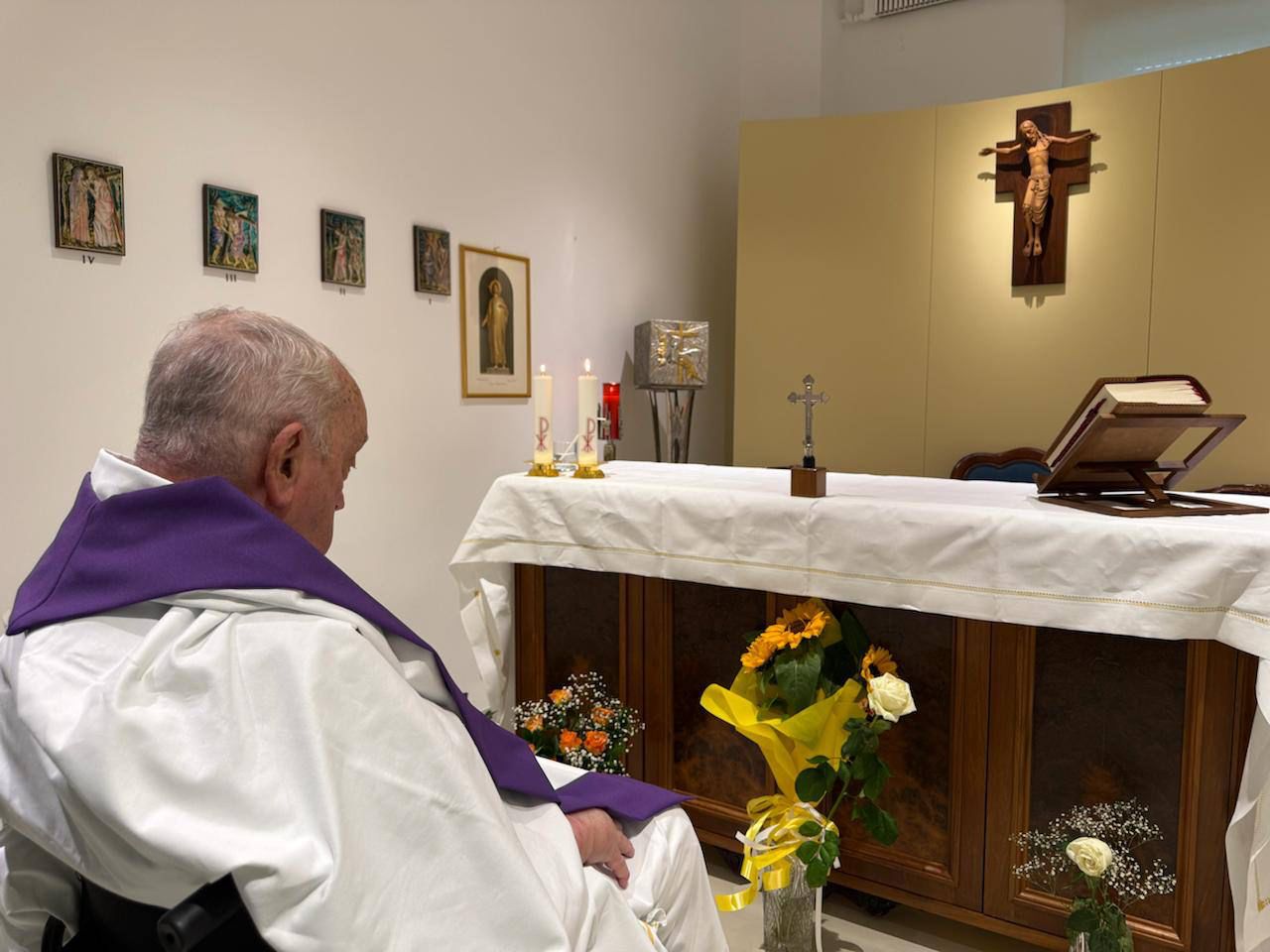 Papa Francisco em momento de oração em um altar, com elementos religiosos ao fundo, incluindo flores, velas e a cruz.