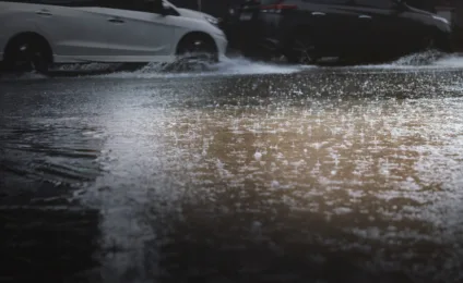 Imagem de chuva intensa criando poças em uma rua, com carros passando ao fundo, mostrando as dificuldades do clima adverso.