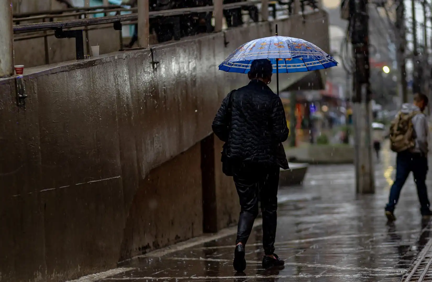 Uma pessoa caminhando sob a chuva com um guarda-chuva azul, em um dia de chuva em Canoas, na região metropolitana.