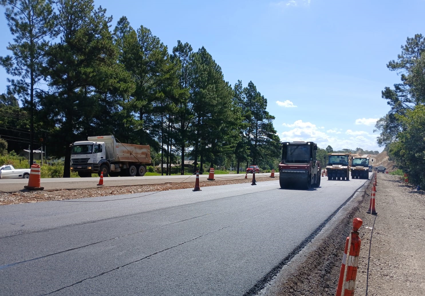 A imagem mostra a duplicação da BR-386, com máquinas pesadas realizando obras e cones de sinalização ao longo da via, cercada por árvores. É um dia ensolarado e as atividades de pavimentação estão em andamento.