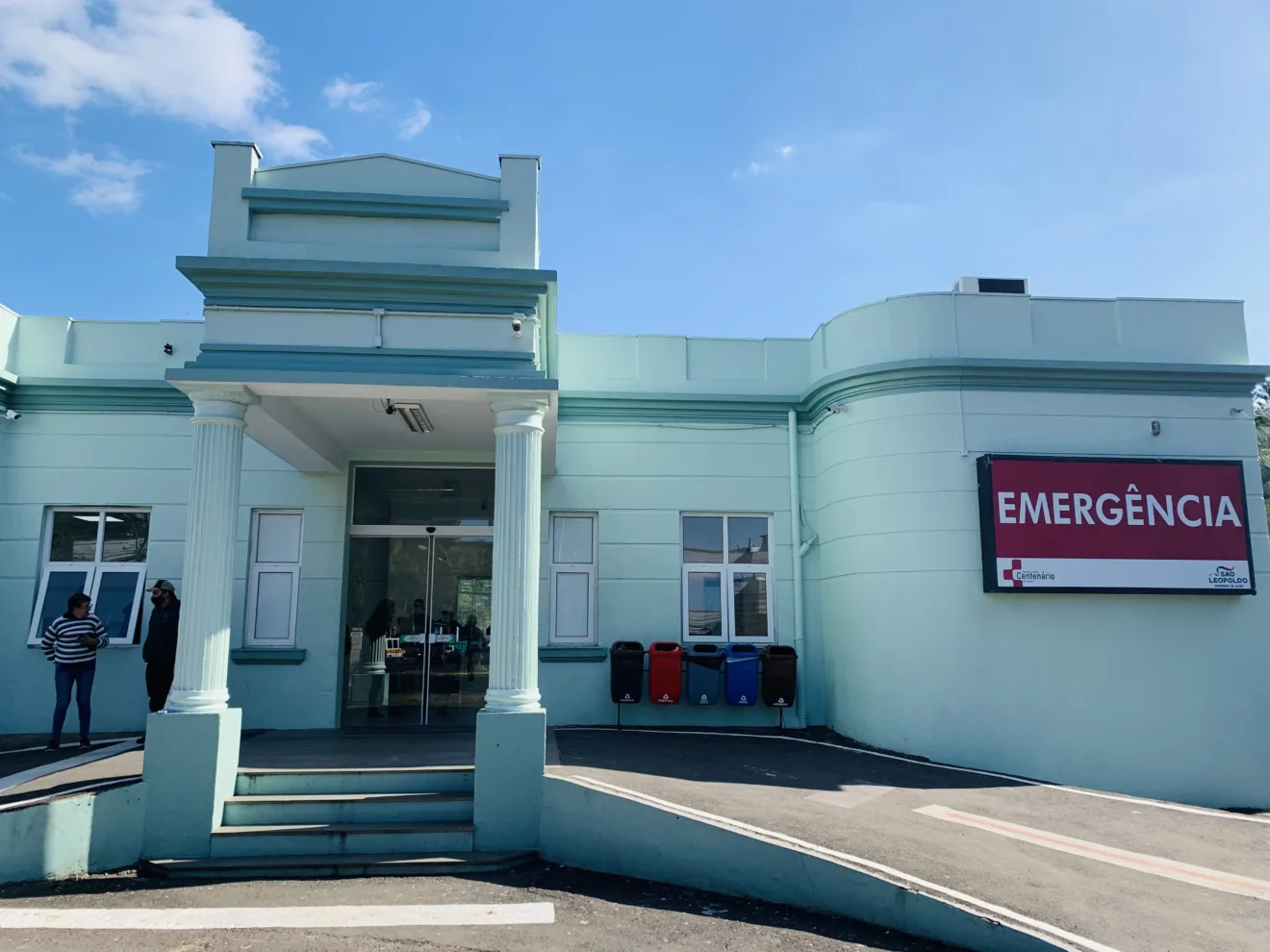 Fachada do Hospital Centenário em São Leopoldo, destacando a entrada do pronto atendimento de emergência, com céu azul ao fundo.