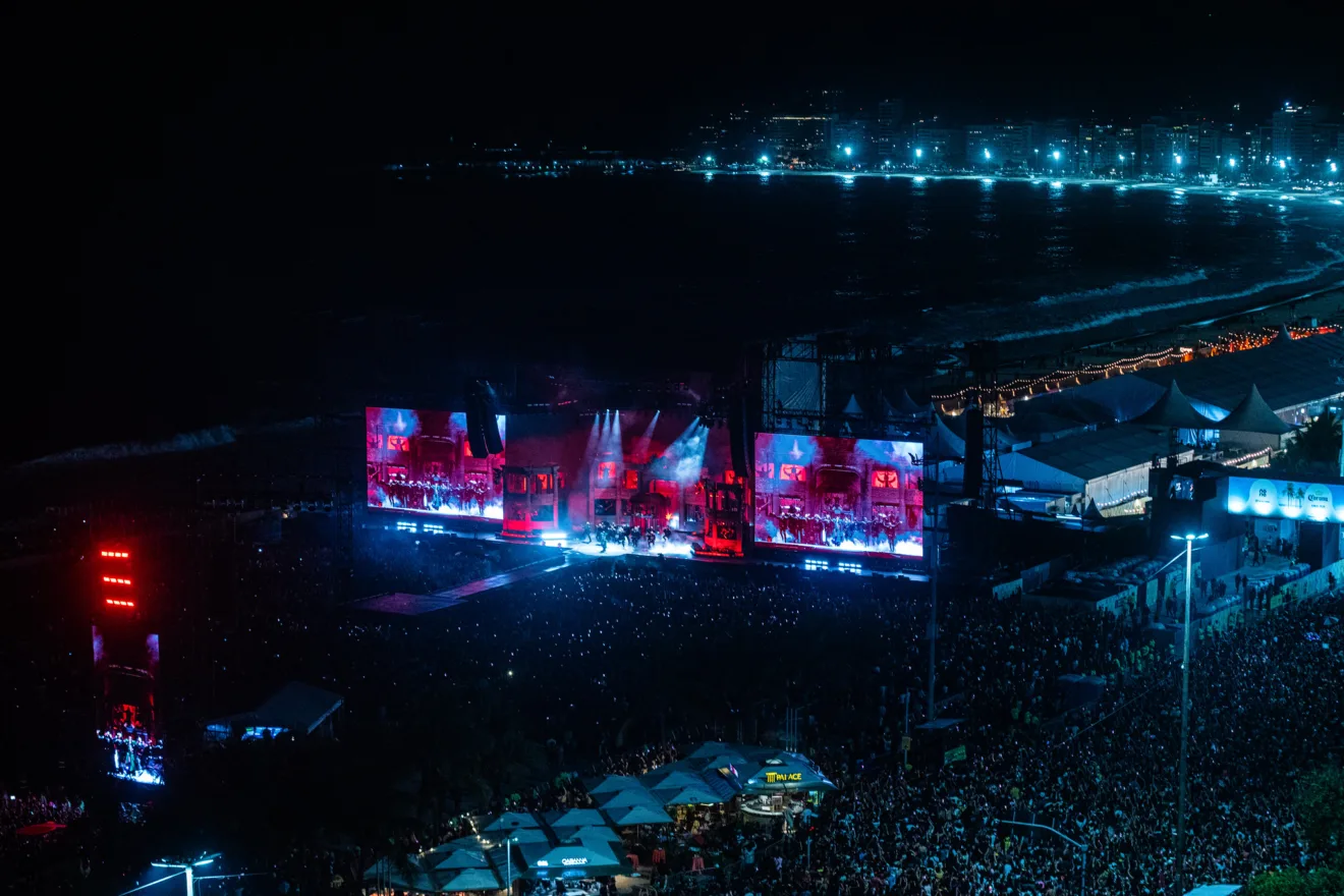 Foto panorâmica do palco do Show da Lady Gaga em Copacabana. É noite e há luzes coloridas e milhares de pessoas
