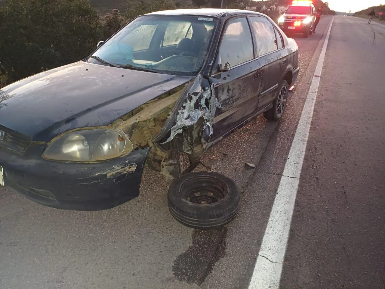 Um carro de cor escura com marca de acidente em meio a uma rodovia