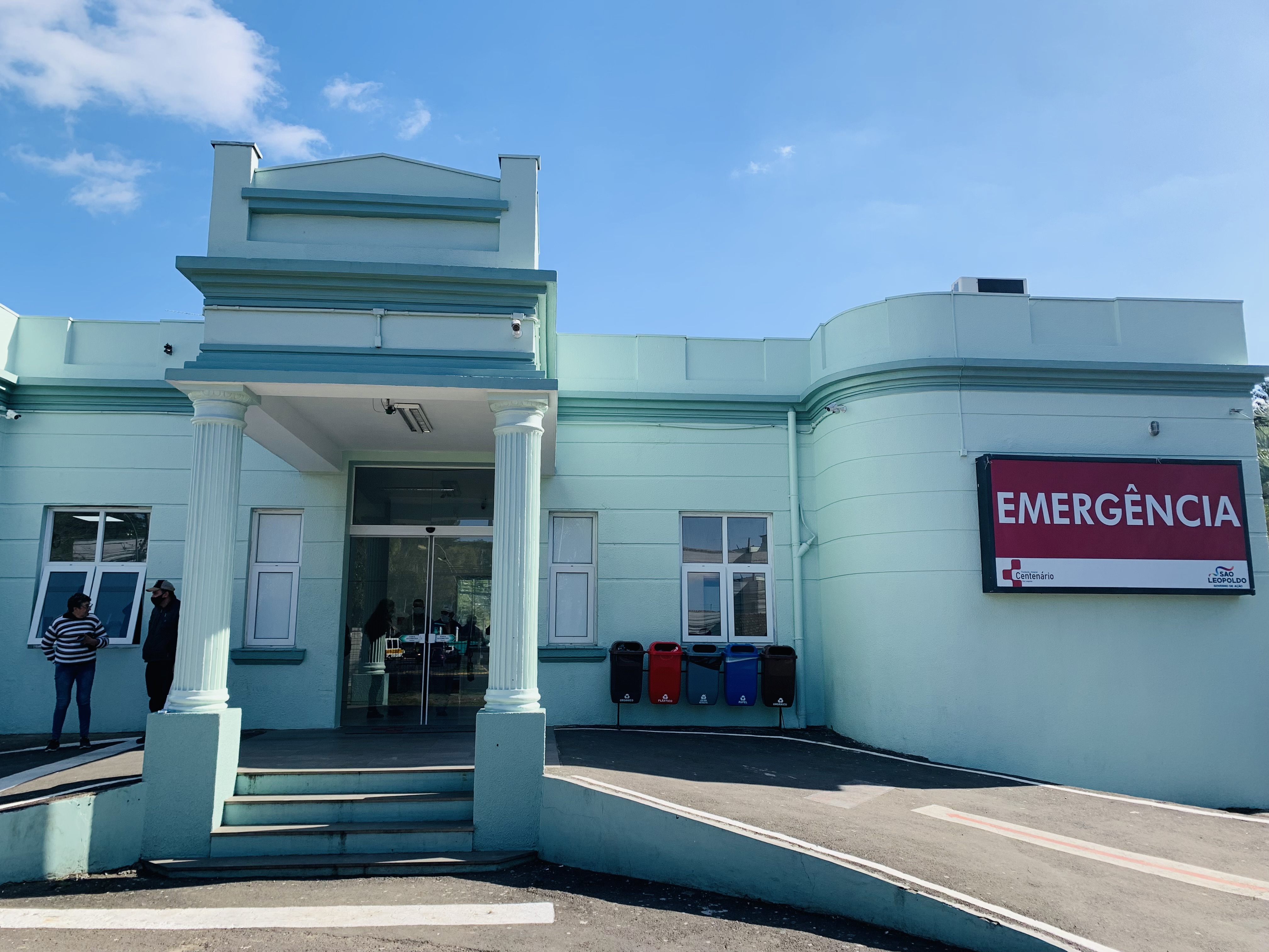 Fachada do Hospital Centenário em São Leopoldo, destacando a entrada do pronto atendimento de emergência, com céu azul ao fundo.