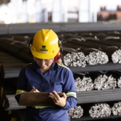Engenheira ou operária de segurança industrial usando capacete amarelo e fone de proteção, verificando inventário de materiais metálicos em ambiente industrial.