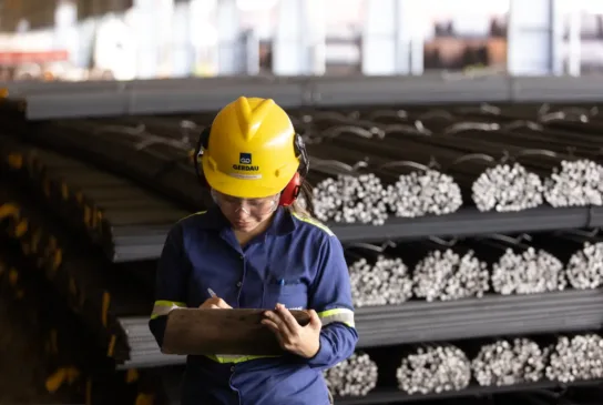 Engenheira ou operária de segurança industrial usando capacete amarelo e fone de proteção, verificando inventário de materiais metálicos em ambiente industrial.